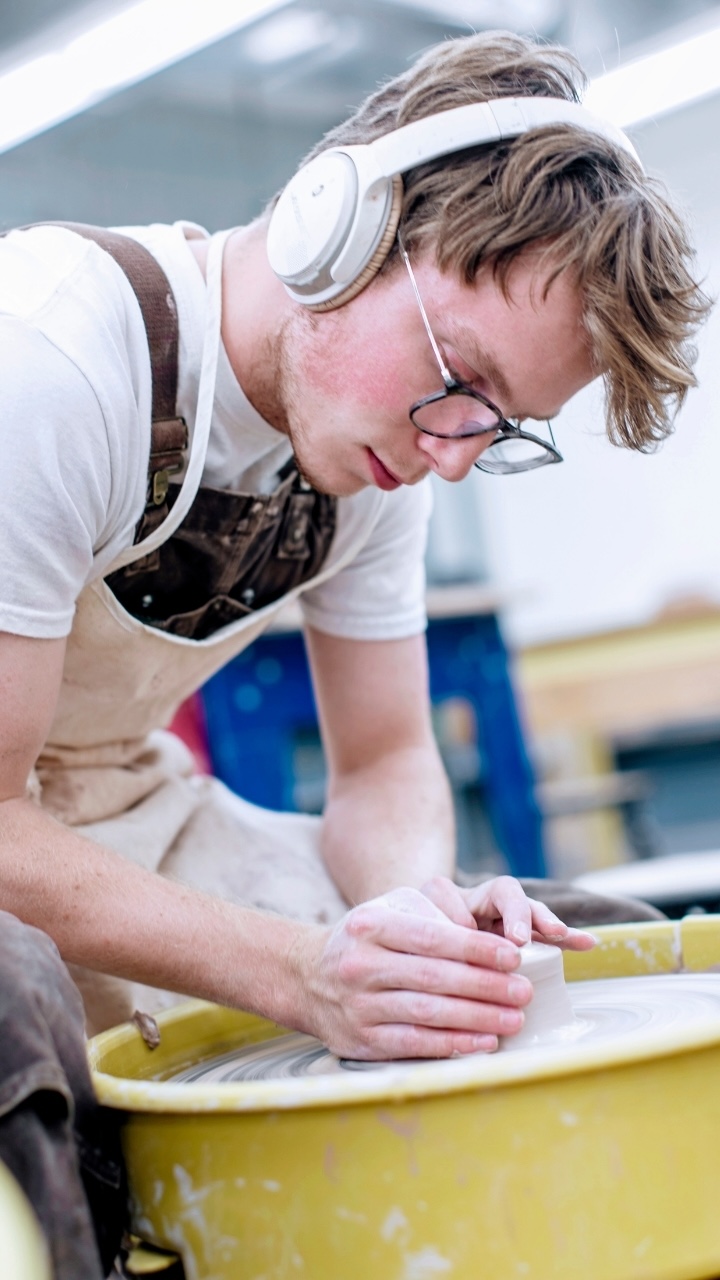 An Allegheny College student molds clay at a pottery wheel with headphones on