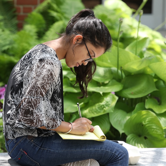 Student sitting on a concrete ledge outside and writing on a yellow notepad