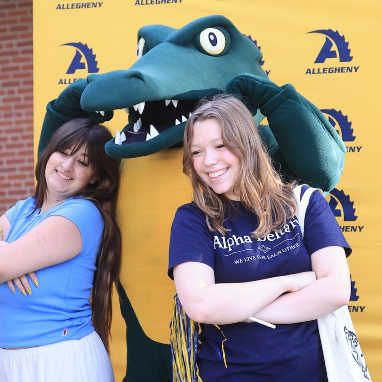 Two Allegheny College students post with the college's alligator mascot, Chompers, in front of a yellow backdrop