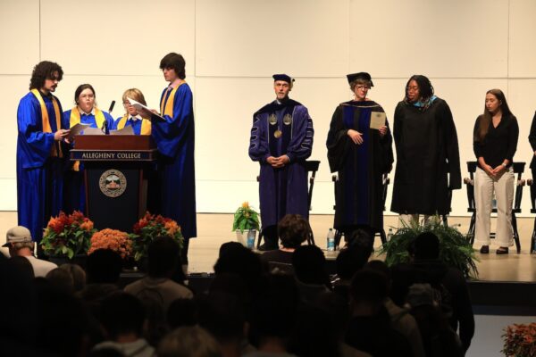 People stand on a stage singing, with a group of four in graduation regalia singing into a microphone on a podium