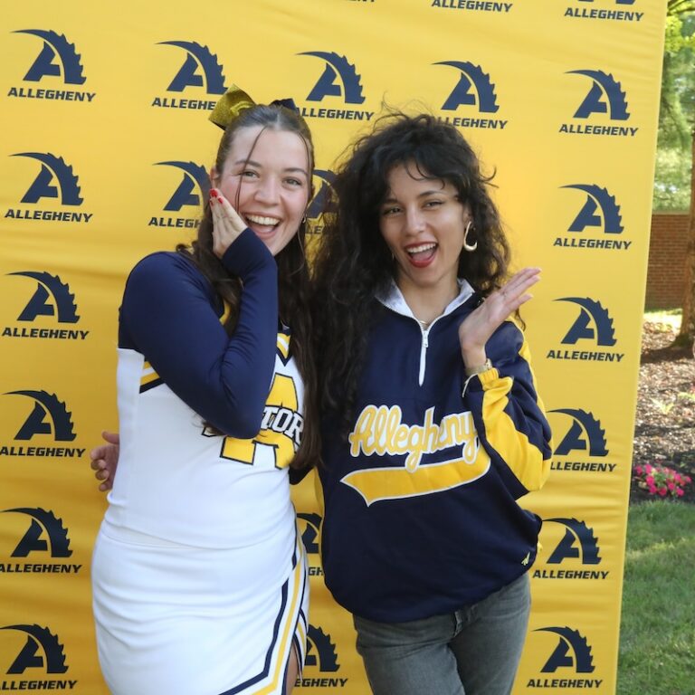 Two Allegheny College students stand in front of a yellow banner with the Allegheny Athletics logo behind them