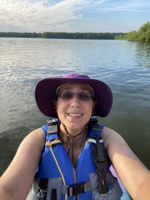 a woman wearing a hat and sunglasses taking a selfie with a lake in the background