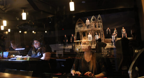 students in an audio booth with the reflection of the stage on the glass in front of them