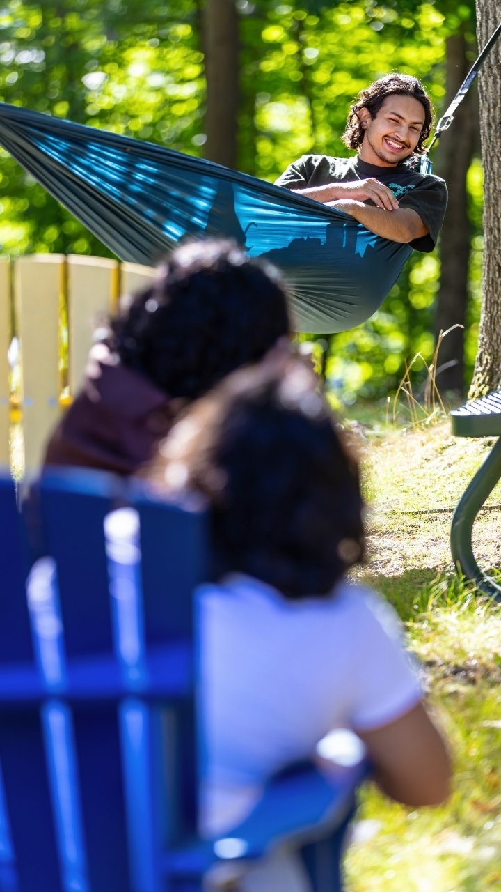 A person swings in a hammock and smiles at friends sitting in adirondack chairs