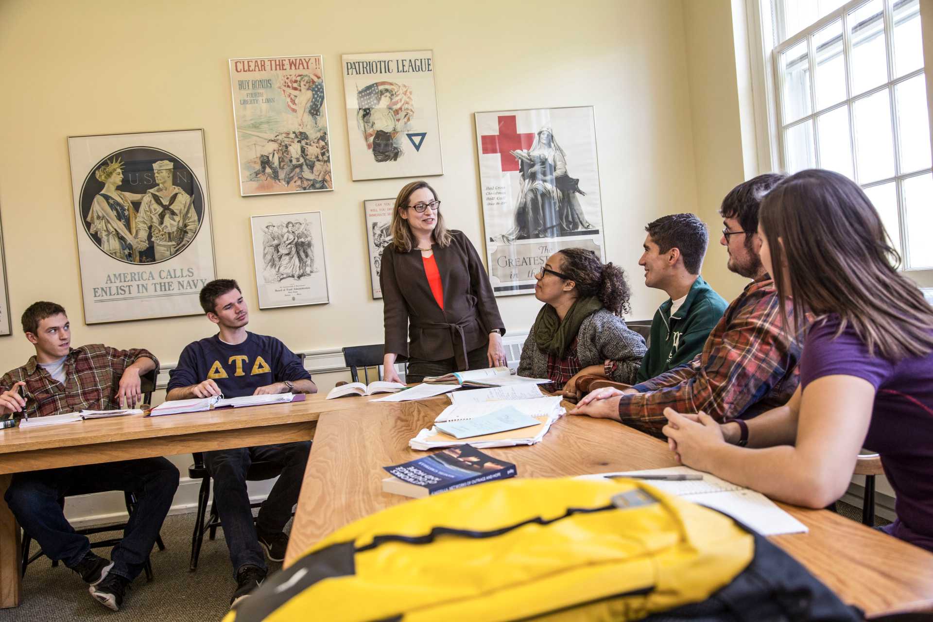 a person standing in front of a group of people in a classroom with a yellow backpack on a desk in the foreground