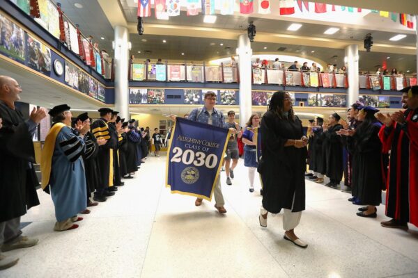 A person walks in a procession holding a banner that reads "Allegheny College Class of 2030" with faculty flanking them