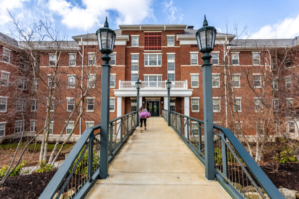 a student walking on a bridge in front of a big brick building with a white balcony