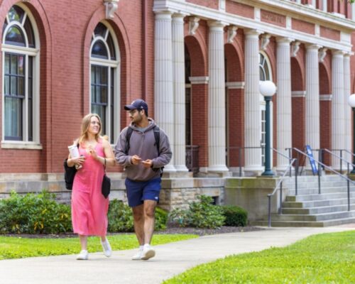 Two Allegheny College students walk down a sidewalk in front of a red brick neoclassical building