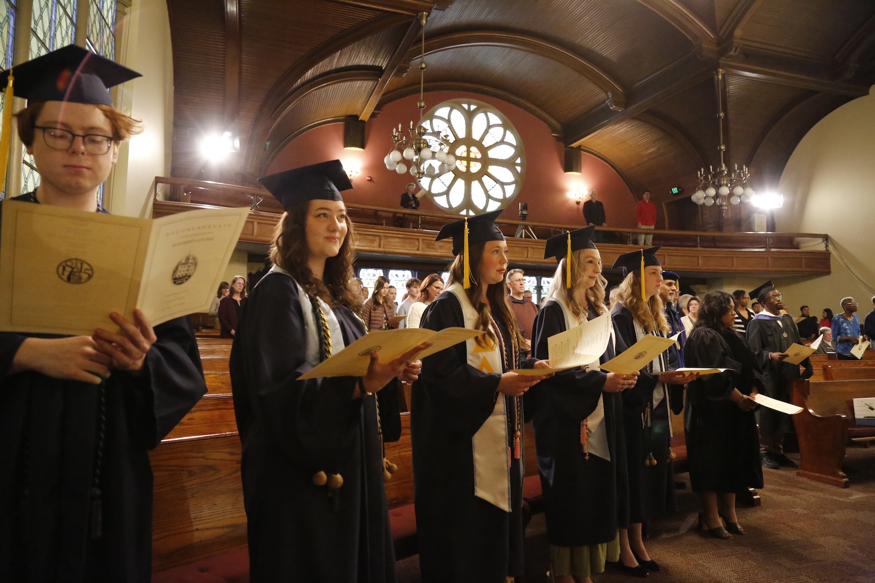 College students wearing graduation regalia hold pamphlets and sing inside a chapel