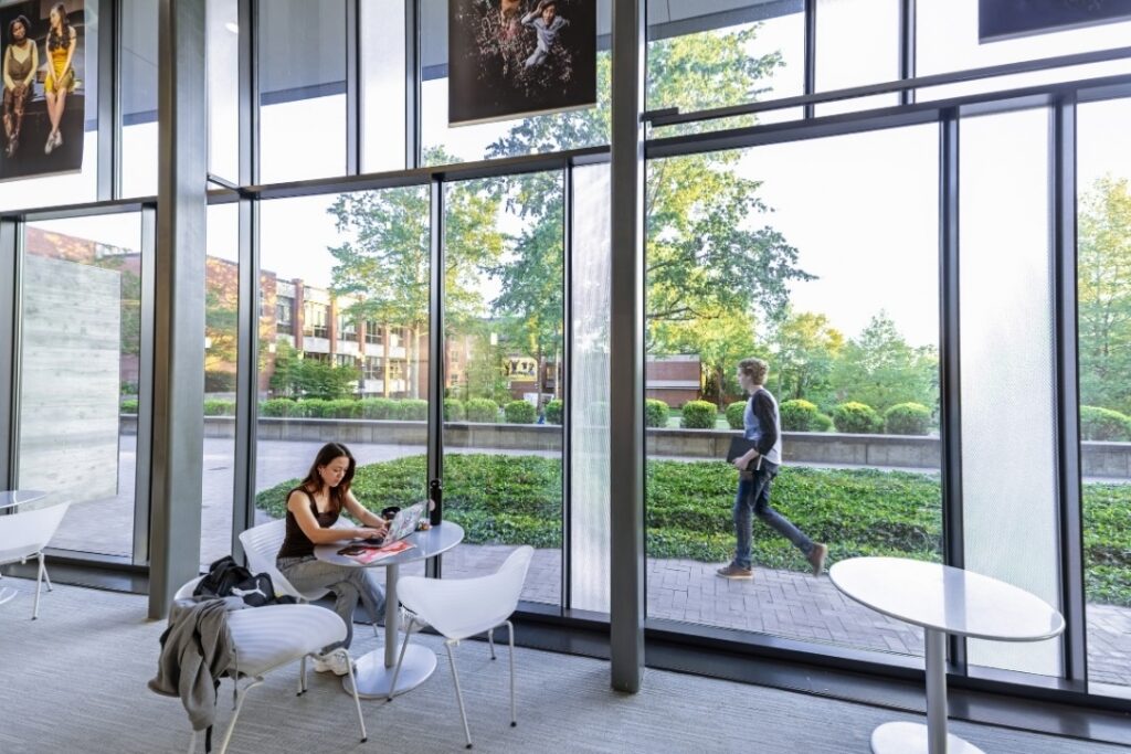 An Allegheny College student works on a laptop with another student walking in the background