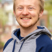 Man with short blonde hair wearing a gray and navy blue hooded sweatshirt