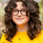 Smiling woman with curly brown hair and glasses wearing a yellow t shirt