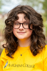 Smiling woman with curly brown hair and glasses wearing a yellow t shirt