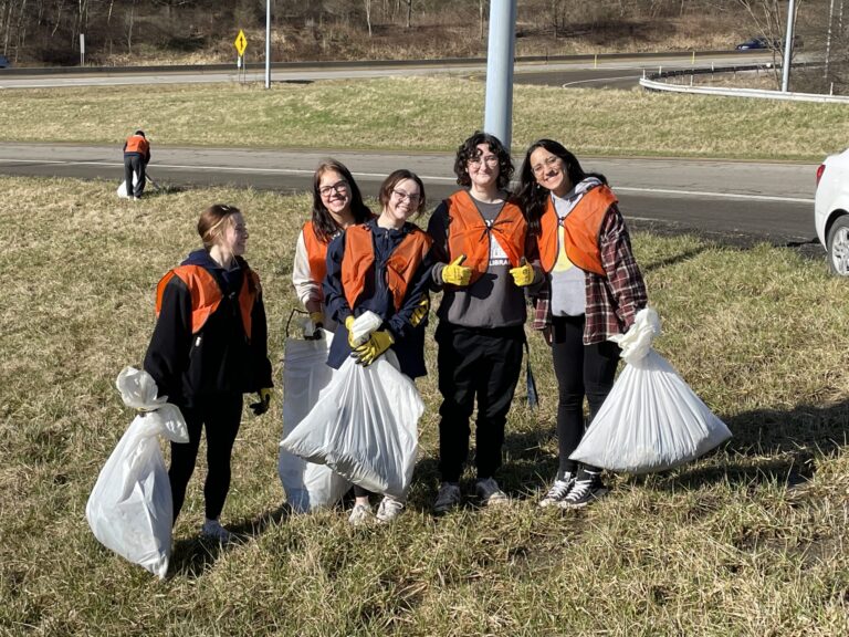 a group of students in orange vests holding plastic bags by the side of a road