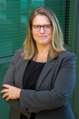 a woman with glasses and a grey jacket posed against a green wall
