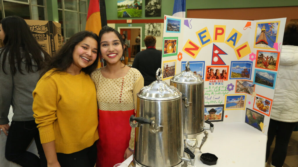 two women standing next to a table with silver containers in front of a sign that reads Nepal
